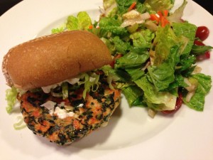 Spinach and feta salmon burger served on a sesame wheat bun slathered in mayo and topped with lettuce.  Romaine, cherry tomato, and carrot salad on the side. (Photo Credit: Adroit Ideals)